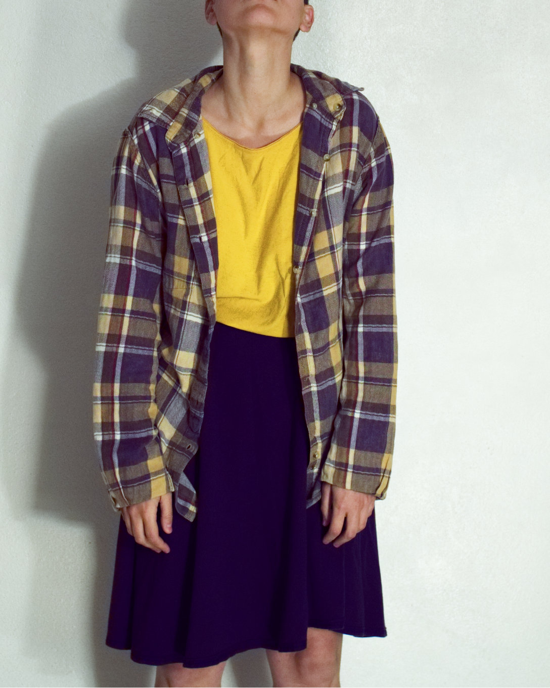 Model wears a yellow and grey plaid shirt, yellow undershirt and dark blue skirt, most of their face is out of the frame, eyes hidden